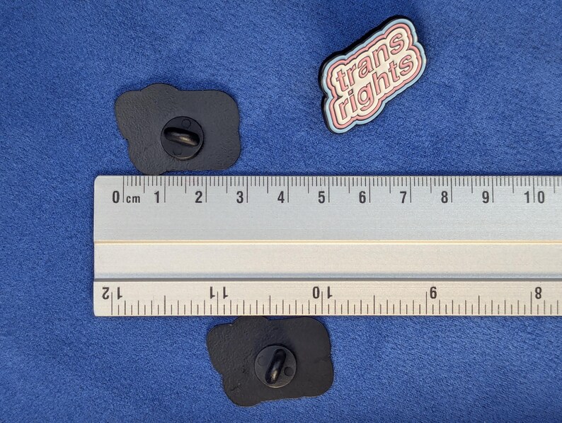 May include: Three enamel pins on a blue surface. One pin reads "trans rights" in pink and white lettering with a blue outline. Two black pins are also visible. A ruler is placed next to the pins.