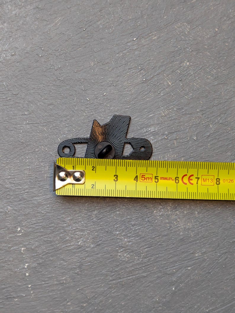 May include: A black metal decorative pin in the shape of a hat, with a pin back. The pin is approximately 1.5 inches long. A yellow Stanley measuring tape is next to the pin.