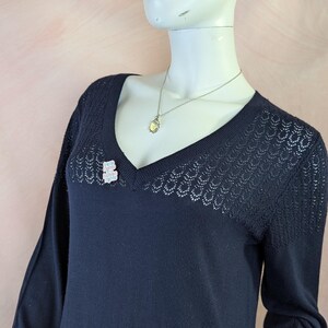 Op de afbeelding: Een marineblauwe trui met lange mouwen, een V-hals en delicate openwerkdetails. Een kleine, sprankelende berenbroche is bij de halslijn gespeld en een zilveren ketting met een hanger hangt.