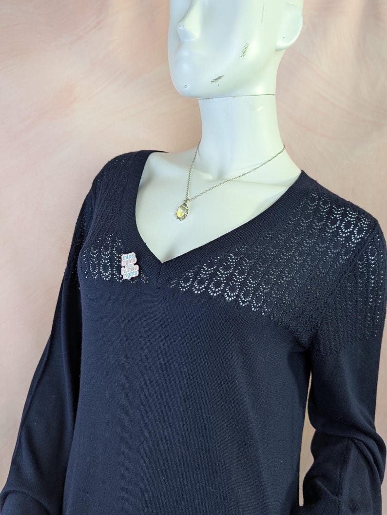 Op de afbeelding: Een marineblauwe trui met lange mouwen, een V-hals en delicate openwerkdetails. Een kleine, sprankelende berenbroche is bij de halslijn gespeld en een zilveren ketting met een hanger hangt.