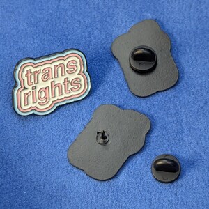 May include: Enamel pin with the text "trans rights" in a white, pink, and blue design. The pin has a black backing with a pin clasp. Two additional black pin backs are also visible.