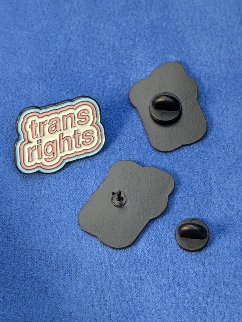 May include: Enamel pin with the text "trans rights" in a white, pink, and blue design. The pin has a black backing with a pin clasp. Two additional black pin backs are also visible.