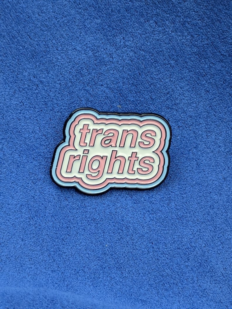May include: A pin with the words "trans rights" in a white, pink, and blue design. The pin has a black border and is set against a blue background.