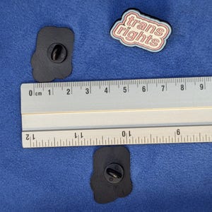 May include: Three enamel pins on a blue surface. One pin reads "trans rights" in a white and pink design. Two black pins with a rounded shape are also visible. A ruler is placed next to the pins.