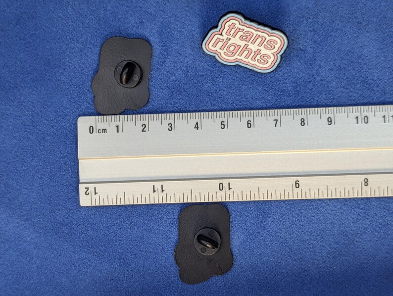 May include: Three enamel pins on a blue surface. One pin reads "trans rights" in a white and pink design. Two black pins with a rounded shape are also visible. A ruler is placed next to the pins.