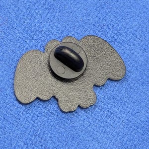 May include: A black enamel pin with a butterfly shape. The pin has a black clutch back and a textured surface. The pin is set against a blue fabric background.