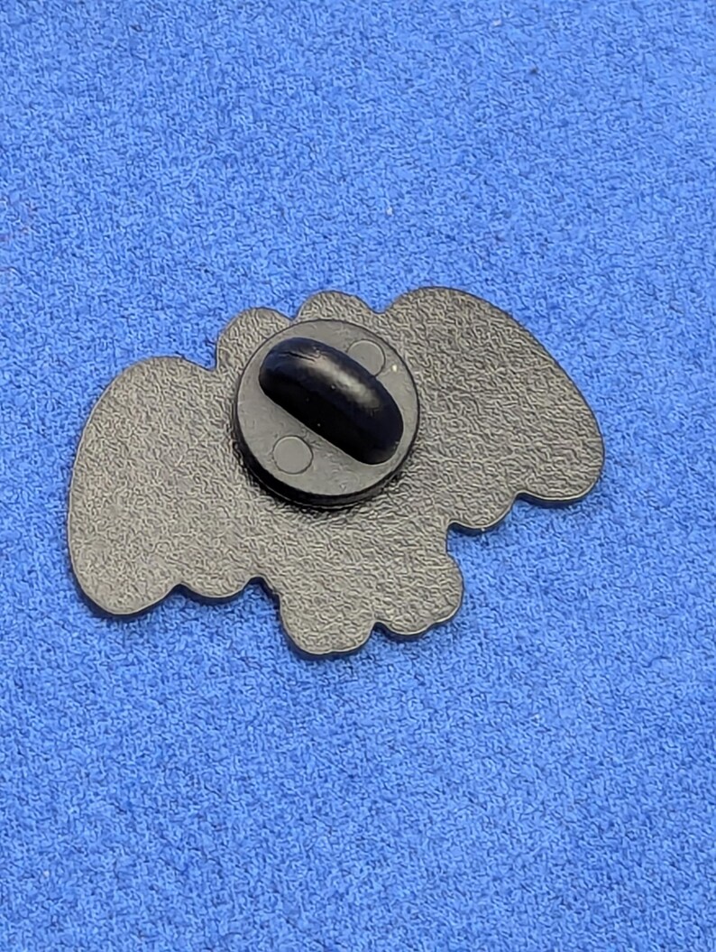 May include: A black enamel pin with a butterfly shape. The pin has a black clutch back and a textured surface. The pin is set against a blue fabric background.