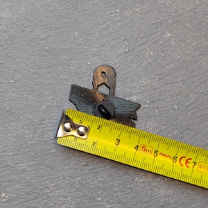May include: A black enamel pin with a sunburst design, approximately 1.5 inches wide, is measured with a yellow tape measure. The pin has a butterfly clutch backing. The background is a textured gray surface.