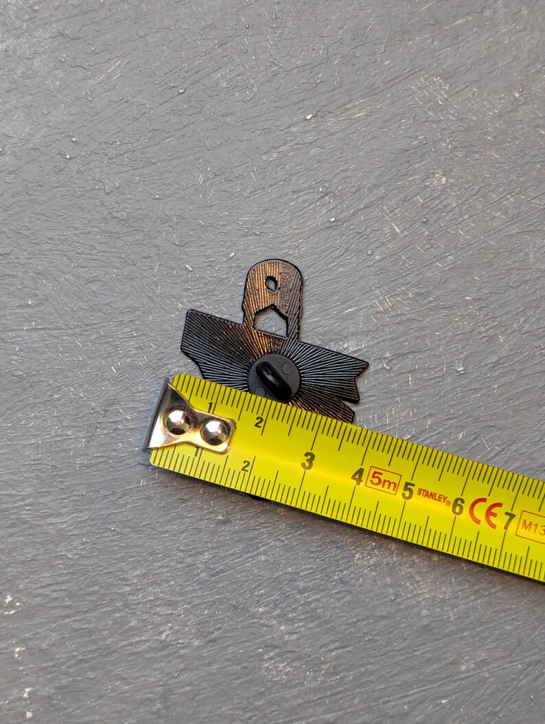 May include: A black enamel pin with a sunburst design, approximately 1.5 inches wide, is measured with a yellow tape measure. The pin has a butterfly clutch backing. The background is a textured gray surface.