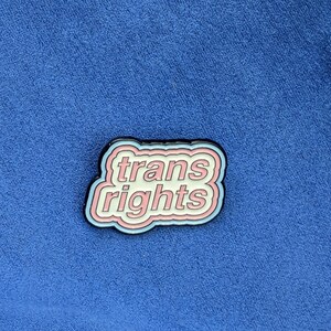 May include: A pin with the words "trans rights" in a white, pink, and blue design. The pin has a black border and is set against a blue background.