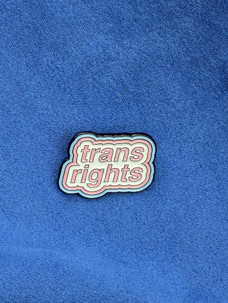 May include: A pin with the words "trans rights" in a white, pink, and blue design. The pin has a black border and is set against a blue background.