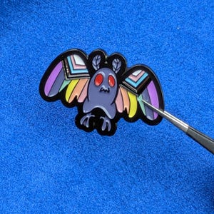 May include: A moth-shaped enamel pin with a black outline. The moth has a purple body, red eyes, and wings with rainbow and pastel color accents. The pin is on a blue surface.