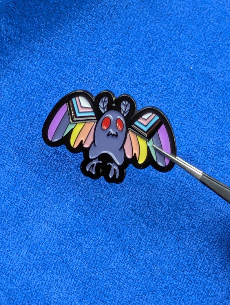 May include: A moth-shaped enamel pin with a black outline. The moth has a purple body, red eyes, and wings with rainbow and pastel color accents. The pin is on a blue surface.