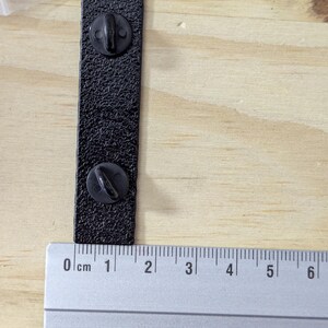 May include: A black rectangular pin with two pin backs, measuring approximately 2.5 inches in length. The pin has a textured surface and is shown next to a ruler for scale. The background is a light wood surface.