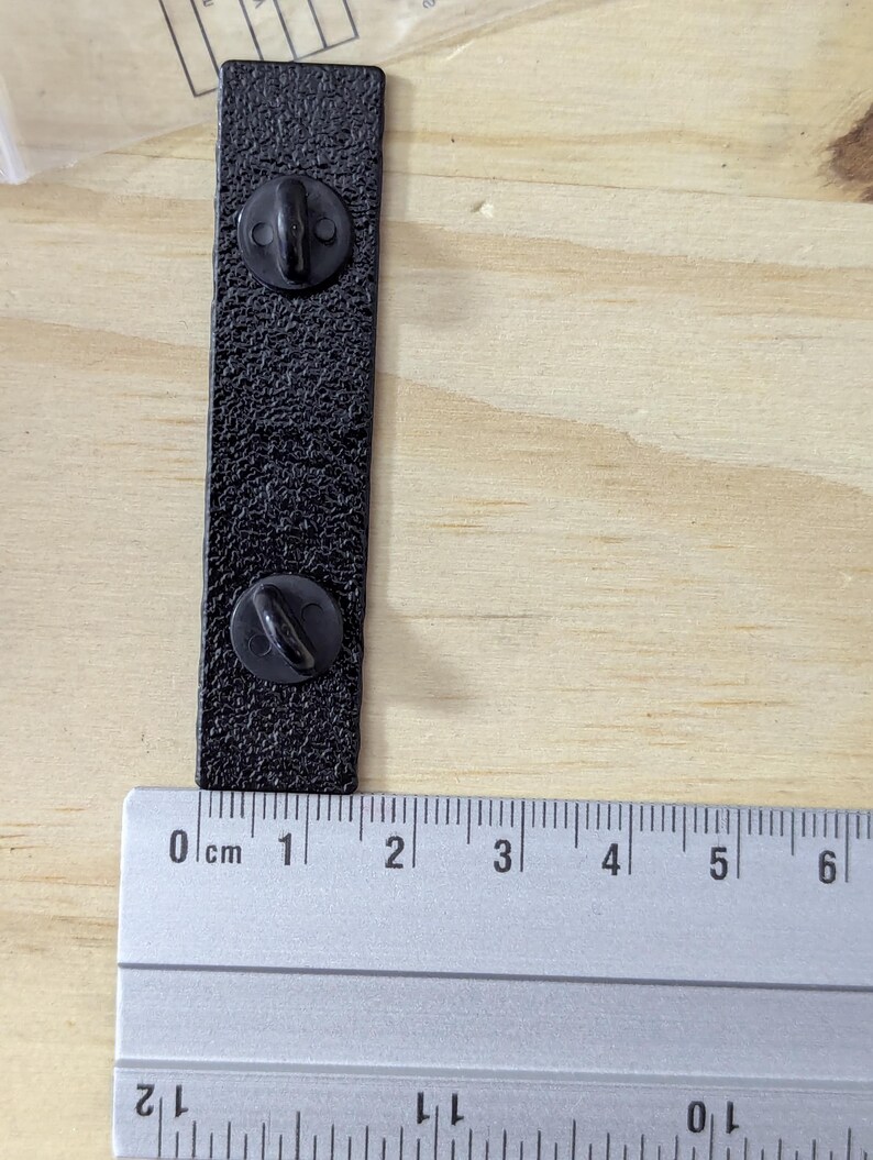 May include: A black rectangular pin with two pin backs, measuring approximately 2.5 inches in length. The pin has a textured surface and is shown next to a ruler for scale. The background is a light wood surface.