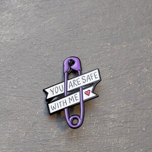 May include: A purple safety pin enamel pin with the words "YOU ARE SAFE WITH ME" on a white banner. A small red heart is next to the words. The pin is on a gray surface.