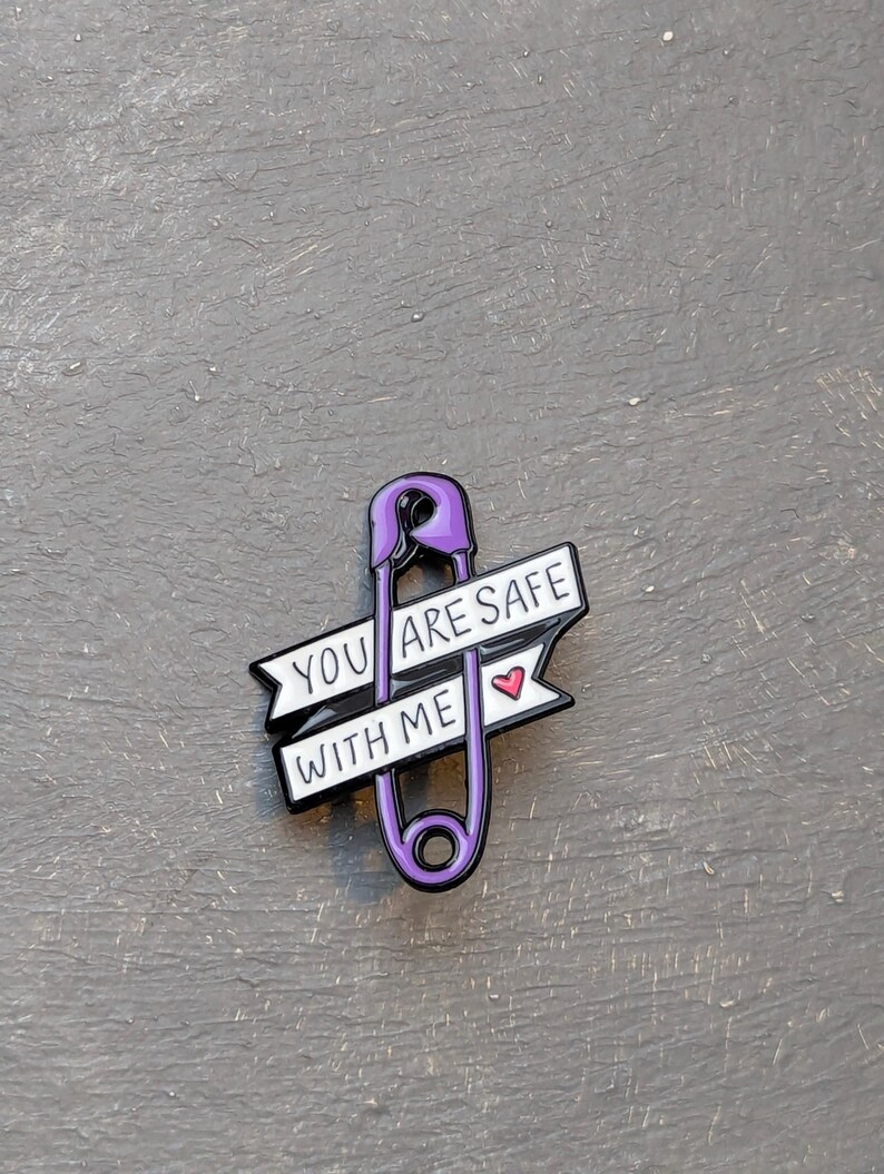 May include: A purple safety pin enamel pin with the words "YOU ARE SAFE WITH ME" on a white banner. A small red heart is next to the words. The pin is on a gray surface.