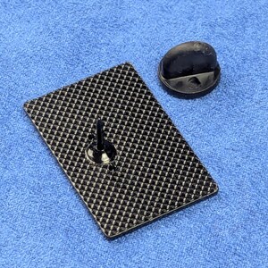 May include: A black rectangular pin with a textured pattern on the back, along with a black pin back. The pin is set against a blue fabric background. The pin's design is simple and functional.