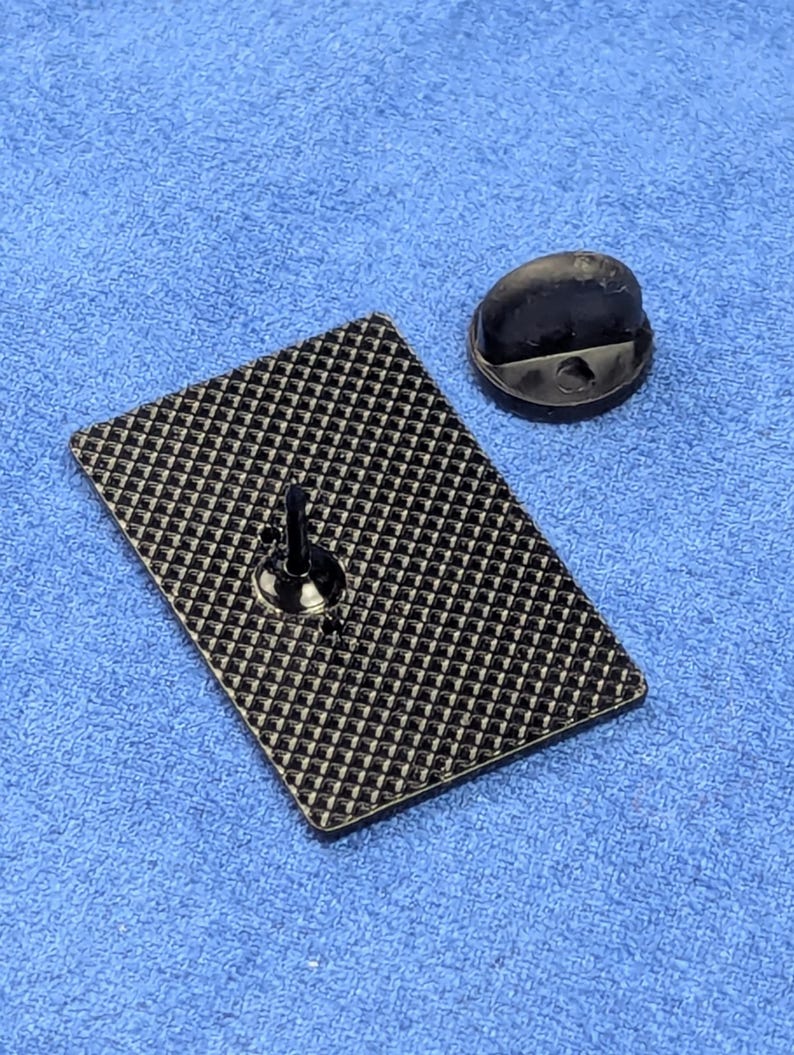May include: A black rectangular pin with a textured pattern on the back, along with a black pin back. The pin is set against a blue fabric background. The pin's design is simple and functional.