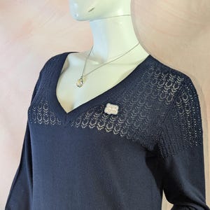 May include: A navy blue sweater with a V-neck and long sleeves. The sweater has a delicate openwork design on the shoulders and neckline. A silver necklace with a pendant and a pin that says "trans rights" are visible.