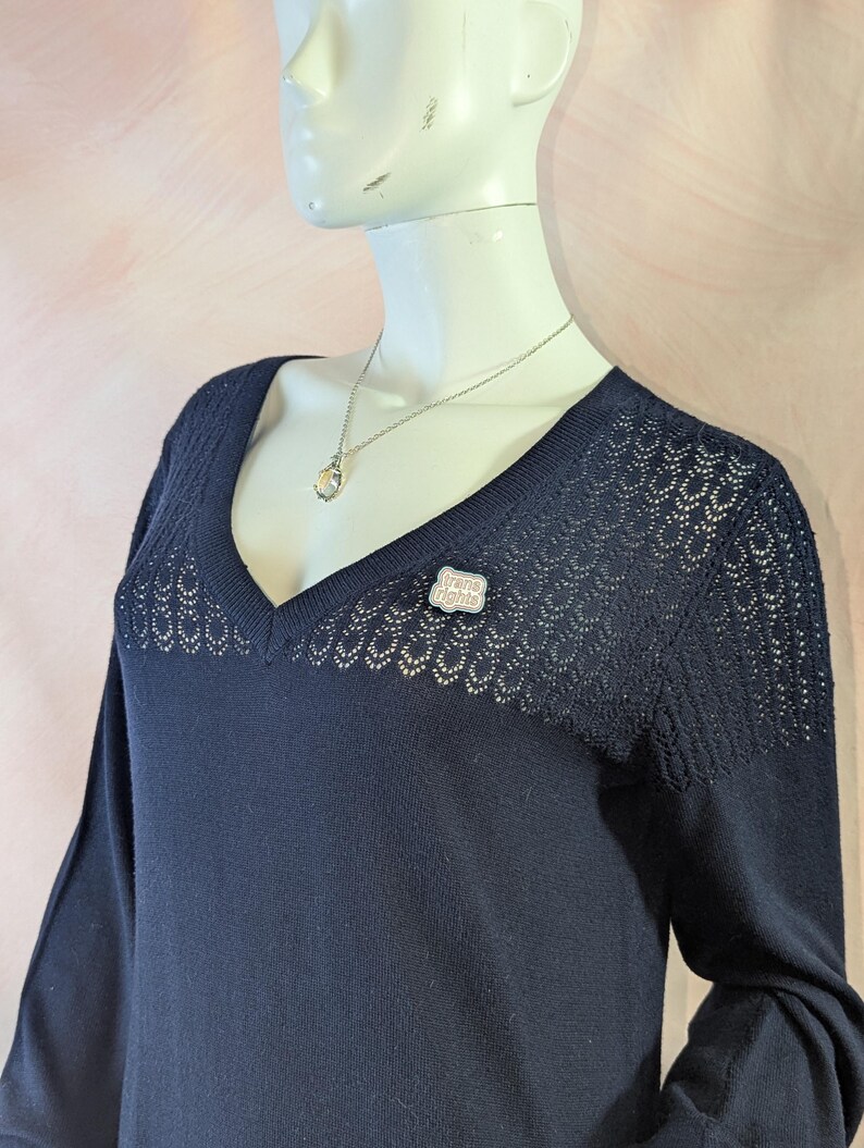 May include: A navy blue sweater with a V-neck and long sleeves. The sweater has a delicate openwork design on the shoulders and neckline. A silver necklace with a pendant and a pin that says "trans rights" are visible.