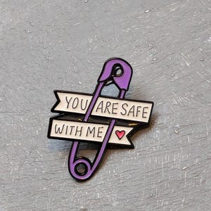 May include: A purple safety pin enamel pin with the text "YOU ARE SAFE WITH ME" in black and white. A small pink heart is on the right side of the text. The pin is on a gray textured surface.
