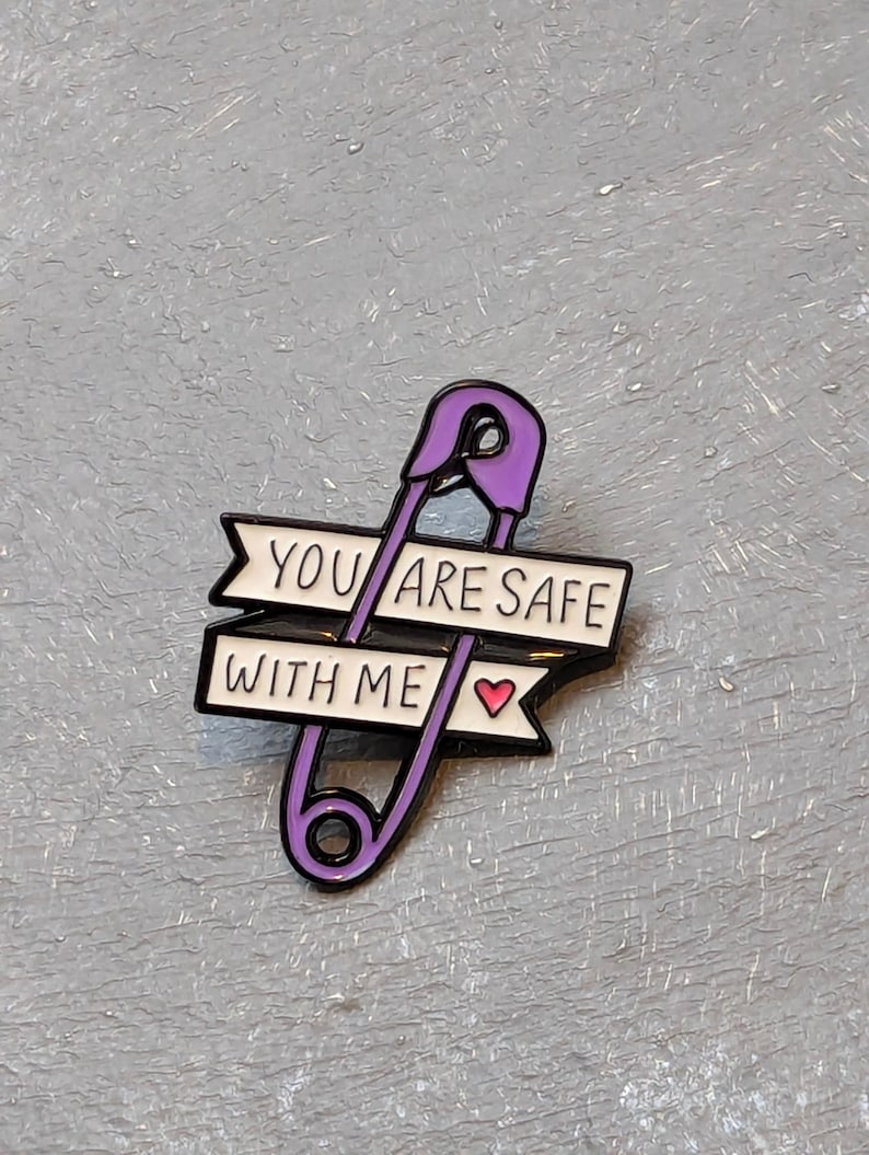 May include: A purple safety pin enamel pin with the text "YOU ARE SAFE WITH ME" in black and white. A small pink heart is on the right side of the text. The pin is on a gray textured surface.