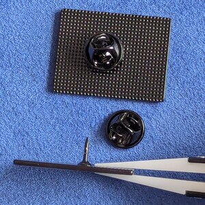 May include: Close-up of the back of a black enamel pin with a square shape and a textured surface. The pin has a black metal clasp. A second black metal clasp is next to the pin. A pair of tweezers holds the pin's needle.