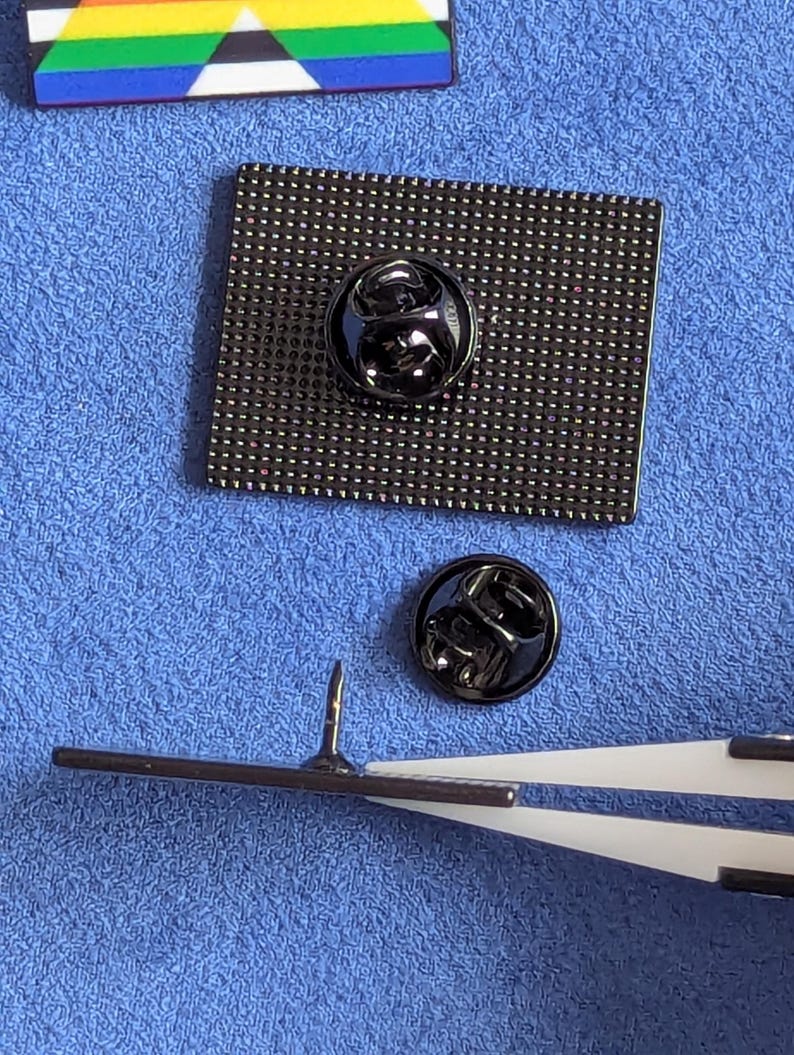 May include: Close-up of the back of a black enamel pin with a square shape and a textured surface. The pin has a black metal clasp. A second black metal clasp is next to the pin. A pair of tweezers holds the pin's needle.