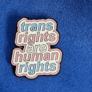 Op de afbeelding: Een rechthoekige emaille pin met de tekst "trans rights are human rights". De pin heeft een witte rand met roze en lichtblauwe tekst op een zwarte achtergrond. De pin is geplaatst op een blauwe stof.