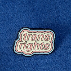 May include: A pin with the words "trans rights" in a white, pink, and light blue design, outlined in black. The pin is set against a blue background.