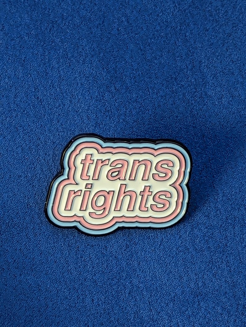 May include: A pin with the words "trans rights" in a white, pink, and light blue design, outlined in black. The pin is set against a blue background.
