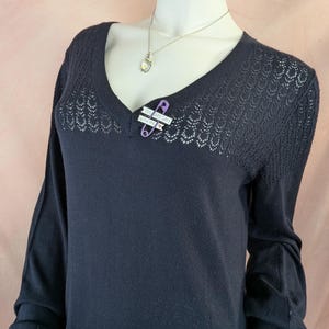 May include: A dark navy blue sweater with a V-neck and long sleeves. The upper chest area has a decorative open knit pattern. A purple safety pin with the text "YOU ARE SAFE WITH ME" is pinned to the sweater. A silver necklace is also visible.