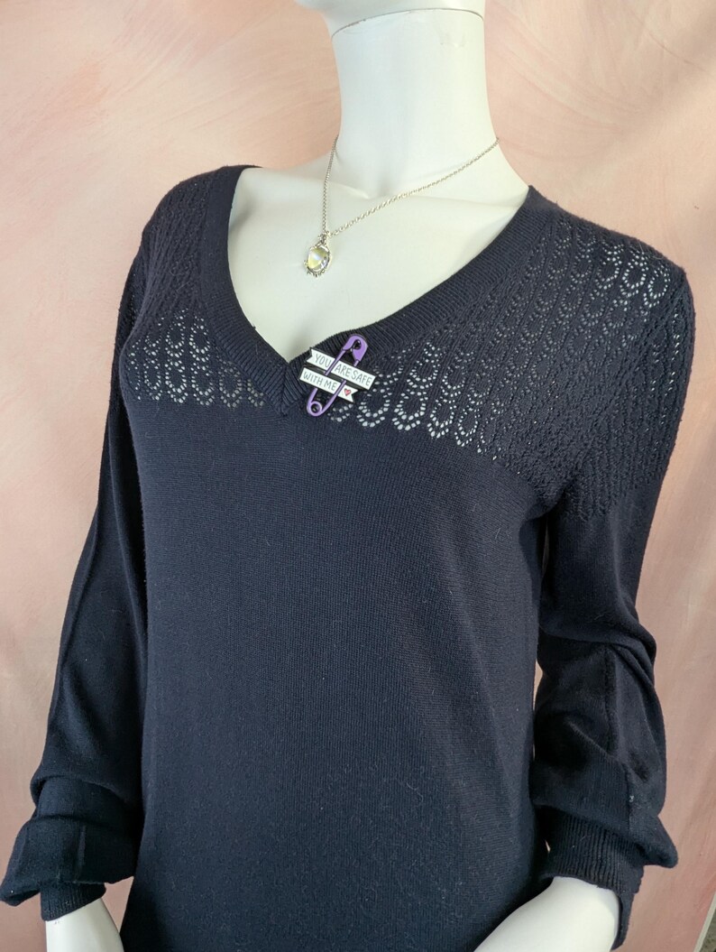 May include: A dark navy blue sweater with a V-neck and long sleeves. The upper chest area has a decorative open knit pattern. A purple safety pin with the text "YOU ARE SAFE WITH ME" is pinned to the sweater. A silver necklace is also visible.
