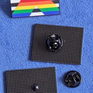 May include: Close-up of three enamel pins. One pin features a rainbow triangle design with black and white stripes. The other two pins are black, rectangular, and have a pin back. One pin back is attached to a pin, and the other is separate.
