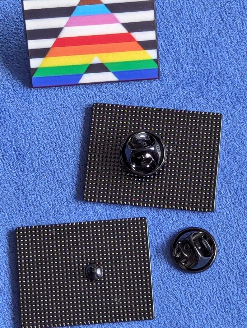 May include: Close-up of three enamel pins. One pin features a rainbow triangle design with black and white stripes. The other two pins are black, rectangular, and have a pin back. One pin back is attached to a pin, and the other is separate.