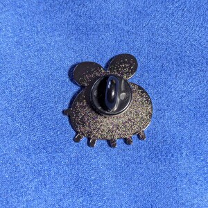 May include: A glittery black enamel pin in the shape of a bee, viewed from the back. The pin has a black post and clasp. The bee's body is round with six small legs. The wings are rounded and the pin is set against a blue background.