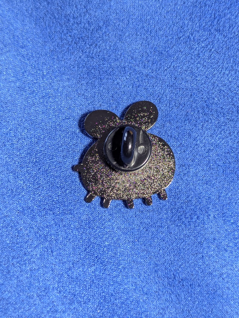 May include: A glittery black enamel pin in the shape of a bee, viewed from the back. The pin has a black post and clasp. The bee's body is round with six small legs. The wings are rounded and the pin is set against a blue background.