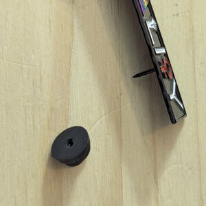 May include: A black rubber bumper and a decorative metal object with a nail are mounted on a light-colored wooden surface. The metal object has a red and silver design with a rainbow-colored stripe.