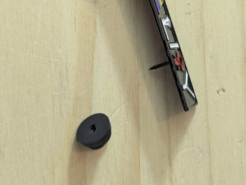 May include: A black rubber bumper and a decorative metal object with a nail are mounted on a light-colored wooden surface. The metal object has a red and silver design with a rainbow-colored stripe.