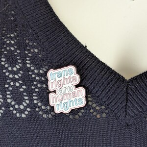 Op de afbeelding: Speld met de tekst "trans rights are human rights" in roze, blauw en wit. De speld is bevestigd op een donkerblauwe gebreide top met een decoratieve kanten rand.