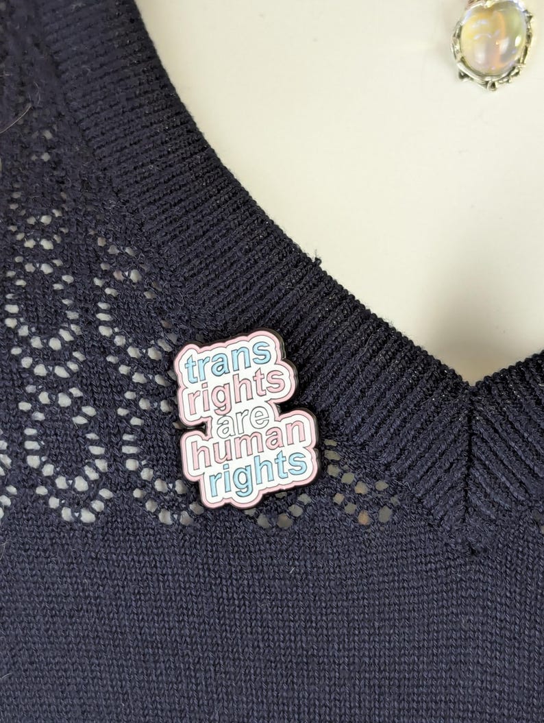 Op de afbeelding: Speld met de tekst "trans rights are human rights" in roze, blauw en wit. De speld is bevestigd op een donkerblauwe gebreide top met een decoratieve kanten rand.