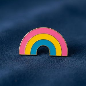 Puede incluir: Pin de esmalte con forma de arcoíris. El pin presenta bandas rosa, amarilla y azul, con un contorno dorado. El pin está sobre un fondo azul oscuro. Accesorio decorativo.