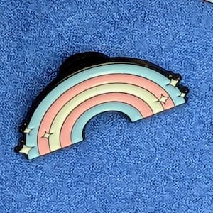 Peut inclure: Une épingle en émail en forme d'arc-en-ciel, avec des rayures rose pastel, jaune et bleu clair. L'arc-en-ciel est souligné en noir et comporte de petits accents d'étoiles à chaque extrémité. L'épingle est posée sur un fond bleu texturé.