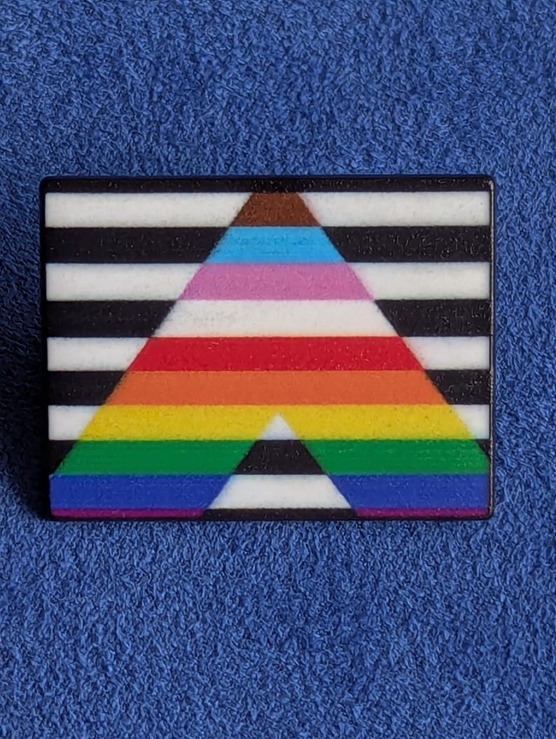 May include: A rectangular pin with a black and white striped background. A colorful triangle is centered on the pin, featuring a rainbow gradient with brown, blue, pink, red, orange, yellow, and green stripes. The pin is set against a blue background.