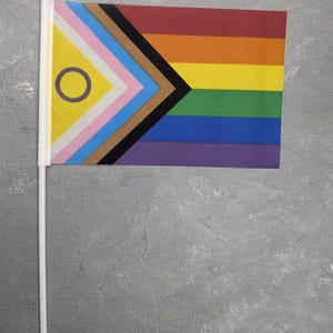 Puede incluir: Una pequeña bandera con un diseño de arcoíris, un triángulo negro y una sección amarilla con un círculo morado. La bandera también incluye secciones rosas, azules y marrones. Está sujeta a un mástil blanco con un adorno dorado.