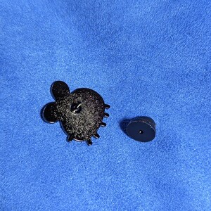 May include: A black enamel pin with a cactus design, featuring a textured surface and a pin back. The pin is shown with its separate black clutch. The background is a blue fabric.