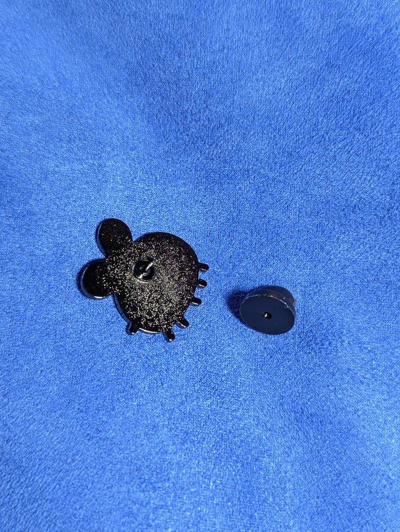 May include: A black enamel pin with a cactus design, featuring a textured surface and a pin back. The pin is shown with its separate black clutch. The background is a blue fabric.