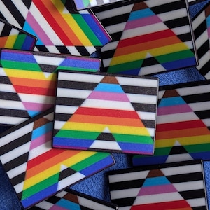 Peut inclure: Plusieurs épingles rectangulaires avec un fond à rayures noires et blanches et un motif triangulaire aux couleurs de l'arc-en-ciel. Les épingles présentent divers schémas de couleurs des drapeaux de la fierté, y compris l'arc-en-ciel traditionnel et les drapeaux LGBTQ+.