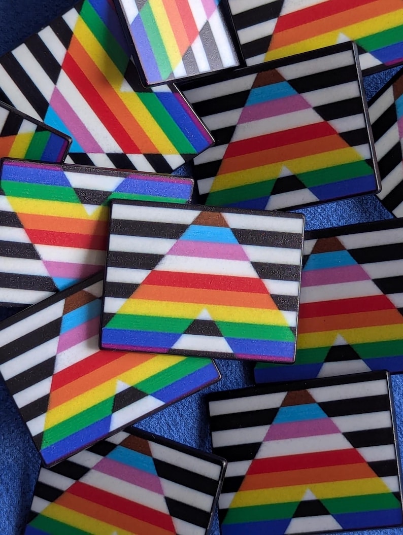 May include: A collection of rectangular pins with a black and white striped background and a rainbow-colored triangle design. The pins feature various pride flag color schemes, including the traditional rainbow, transgender, and other LGBTQ+ flags.